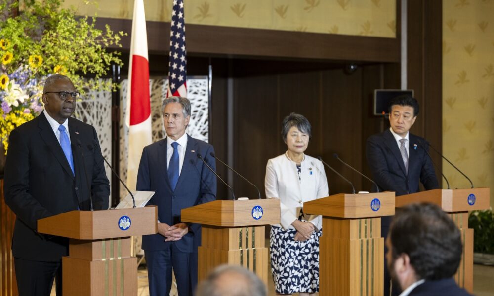 Four individuals stand behind lecterns. Behind them are a Japanese flag and an American flag.