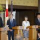 Four individuals stand behind lecterns. Behind them are a Japanese flag and an American flag.