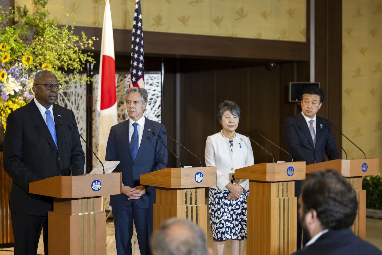 Four individuals stand behind lecterns. Behind them are a Japanese flag and an American flag.